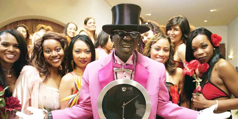   Flavor Flav cilindert visel, rózsaszín öltönyben, és egy csoport nő előtt áll a Flavor of Love egyik jelenetében.