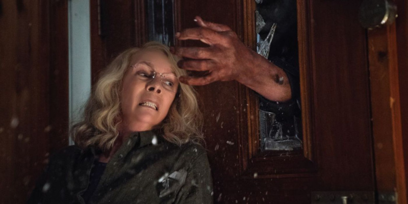   Laurie Strode nekiütközött az ajtónak, miközben egy kéz megpróbálja kinyitni azt egy betört ablakon keresztül 2018 Halloweenkor.